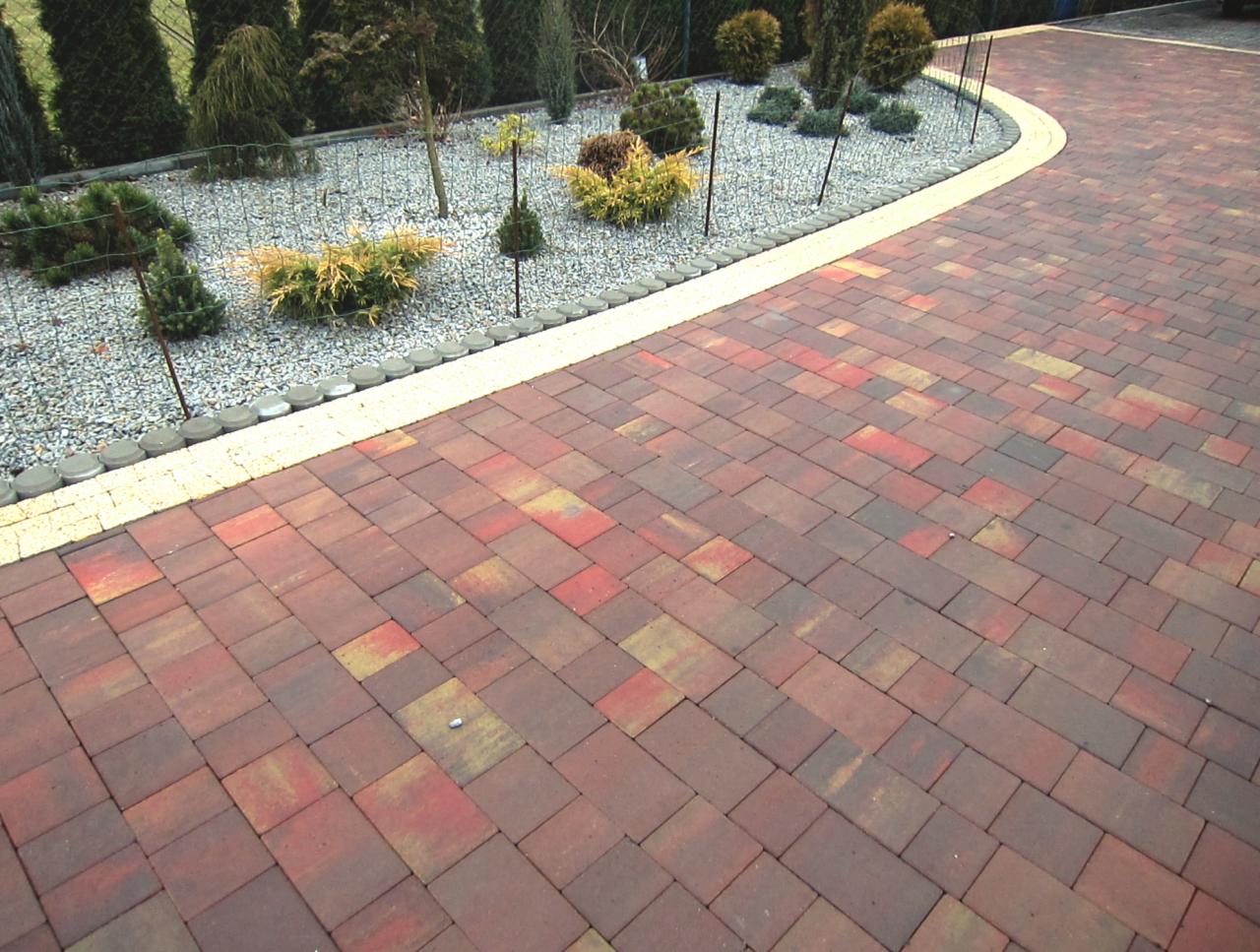 Ułożona, czerwono-brązowa kostka betonowa z obrzeżem w kolorze kremowym, przylegająca do rabaty z szarym żwirem i niskimi roślinami iglastymi.