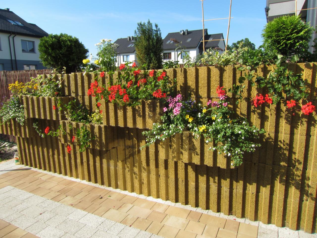 Ozdobny murek z prefabrykatów betonowych, z wnękami wypełnionymi kwitnącymi pelargoniami i innymi kwiatami, tworzący zieloną ścianę w ogrodzie.