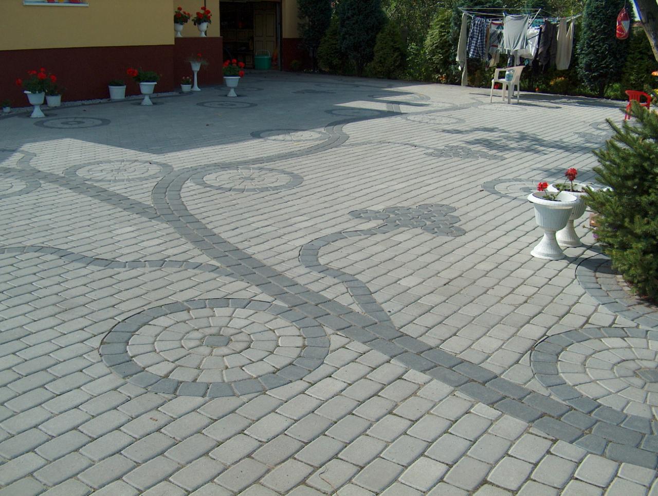 Ułożona kostka betonowa w szarych odcieniach tworząca wzór z okręgów i linii. Widoczne donice z kwiatami, drzewa i suszarka na pranie w tle. Kąt ujęcia z góry.