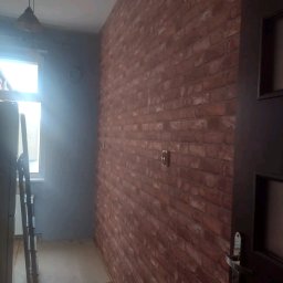 Pawlowolochastyi - Ściana wykończona imitacją czerwonej cegły, z widocznym gniazdkiem elektrycznym i fragmentem ciemnych drzwi, w tle ściana pomalowana na szaro i drabina oparta o ścianę przy oknie.