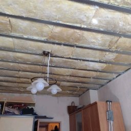 Pawlowolochastyi - Montaż izolacji z wełny mineralnej na suficie podwieszanym z metalową konstrukcją, widoczna lampa wisząca i fragment umeblowania pokoju.