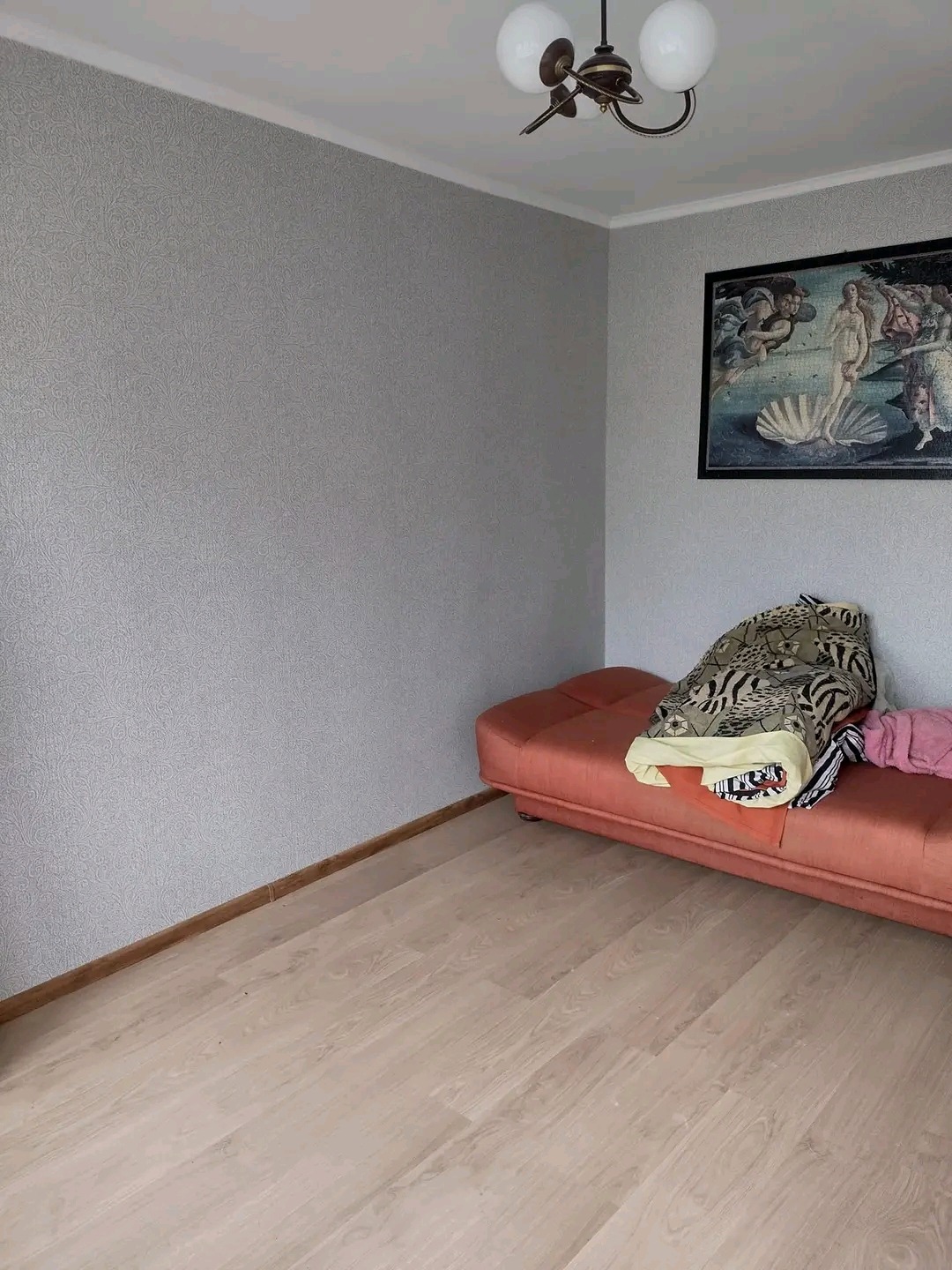 Pokój po remoncie: świeżo położone panele podłogowe, ściany pokryte szarą tapetą ze wzorem, na ścianie wisi reprodukcja obrazu 'Narodziny Wenus', a w rogu stoi pomarańczowa sofa z narzuconą kołdrą.