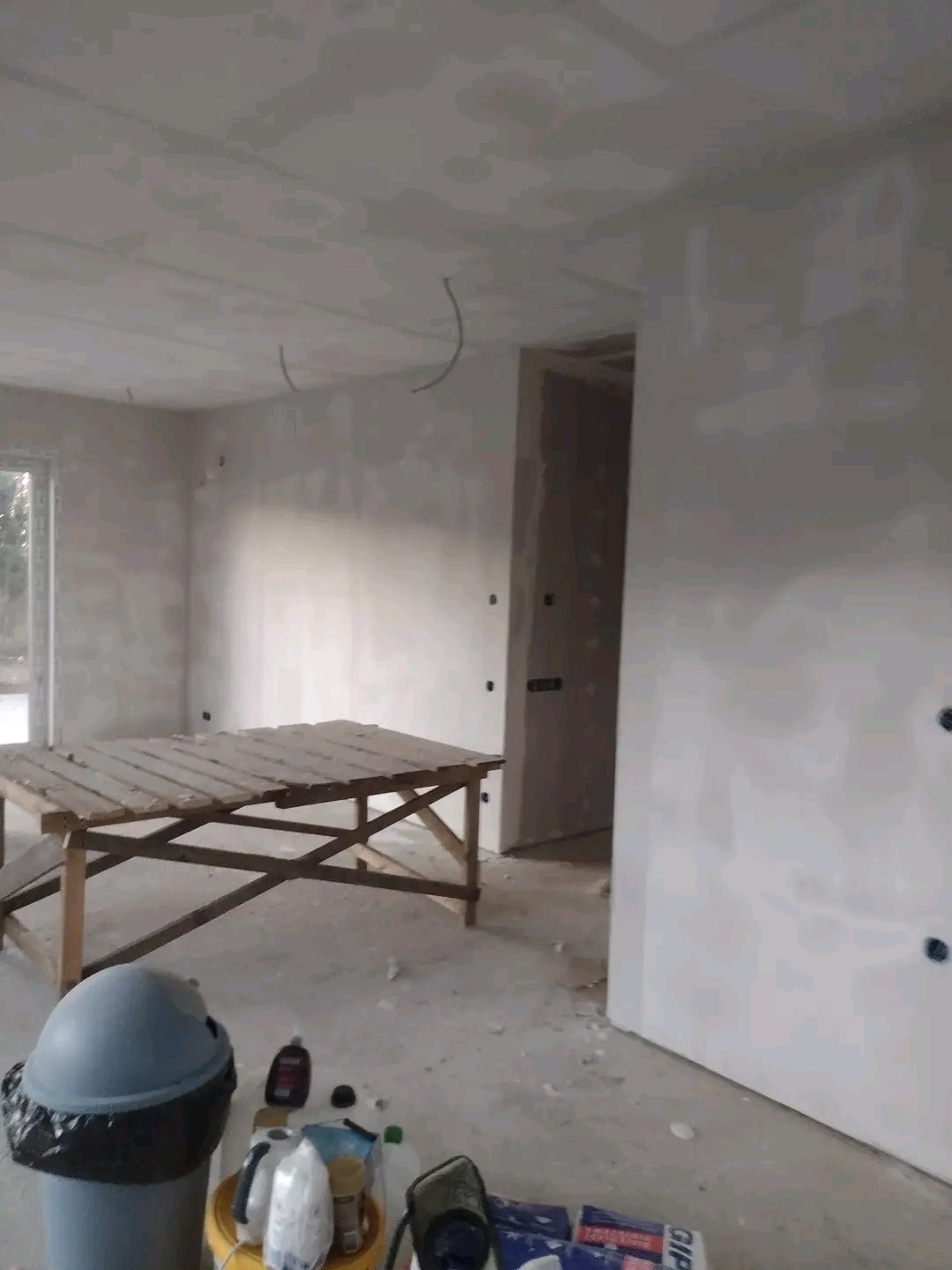 Wnętrze w trakcie prac przygotowawczych do wykończenia ścian: widoczne zagruntowane ściany, drewniany stół roboczy, wiadro z narzędziami i materiałami, wystające przewody elektryczne.