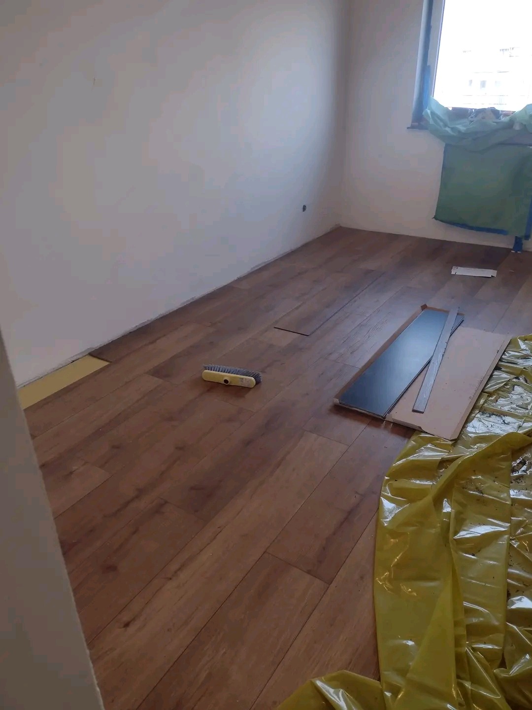 Proces montażu paneli podłogowych w pokoju: widoczne panele, podkład, szczotka do zamiatania, folia ochronna na podłodze, ściany przygotowane do malowania.