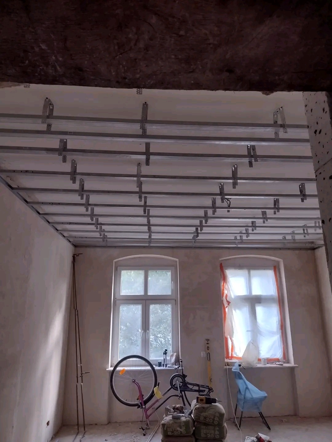 Montaż metalowej konstrukcji sufitu podwieszanego w pomieszczeniu z dwoma oknami, widoczne worki z zaprawą i rower oparty o ścianę.