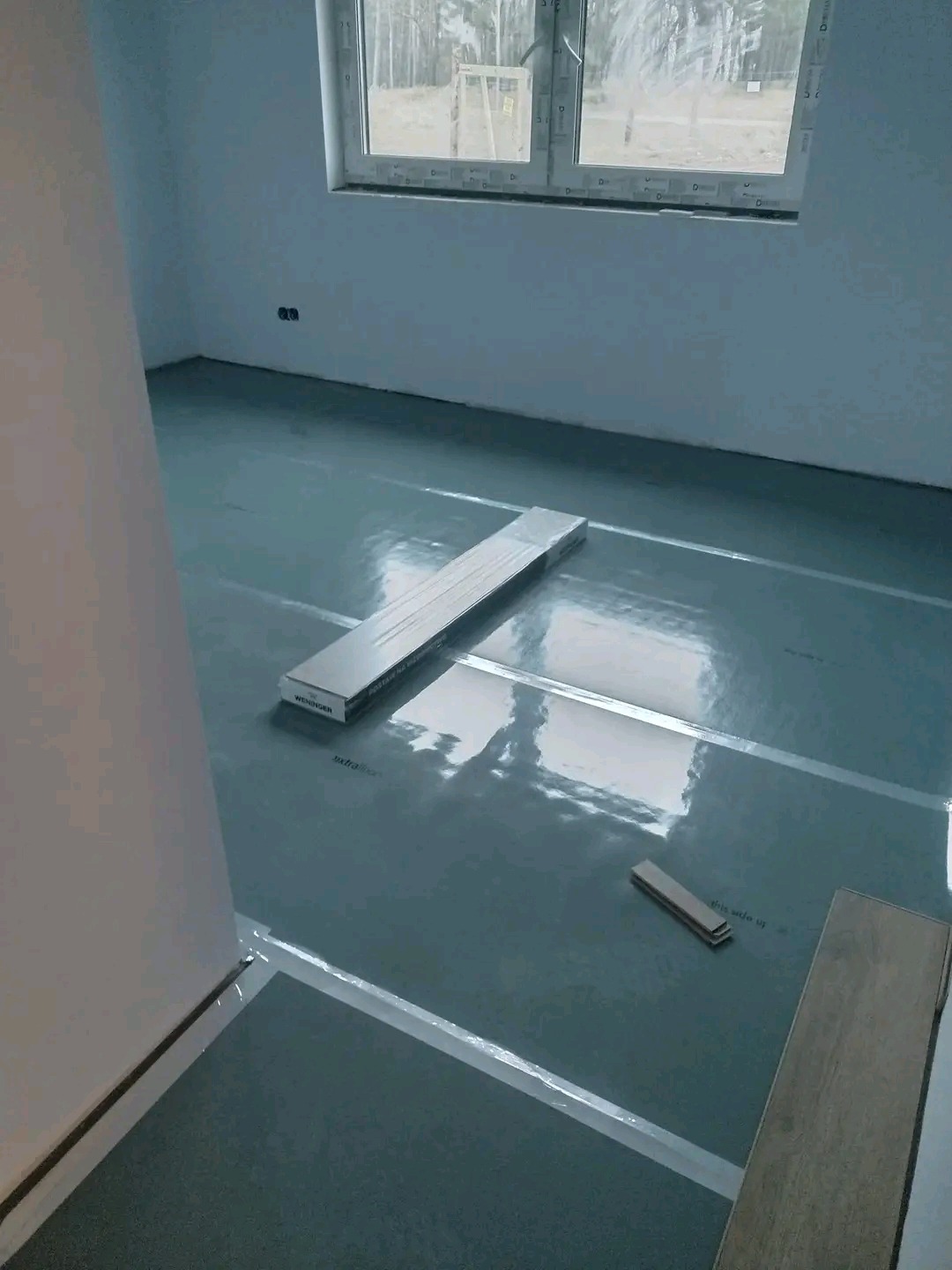 Przygotowanie do ułożenia paneli: rozłożona folia izolacyjna, paczka paneli Weninger i próbka panela na podłodze w jasnym, pustym pokoju z oknem.