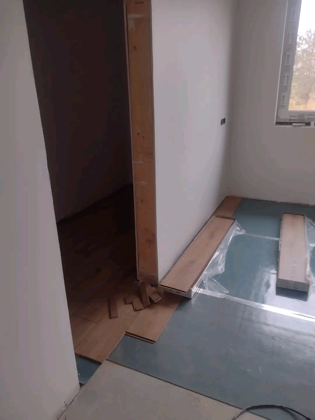 Fragment pokoju w trakcie montażu paneli podłogowych, widoczna folia podkładowa, kilka dociętych elementów paneli przy ościeżnicy, oraz paczka paneli w tle.