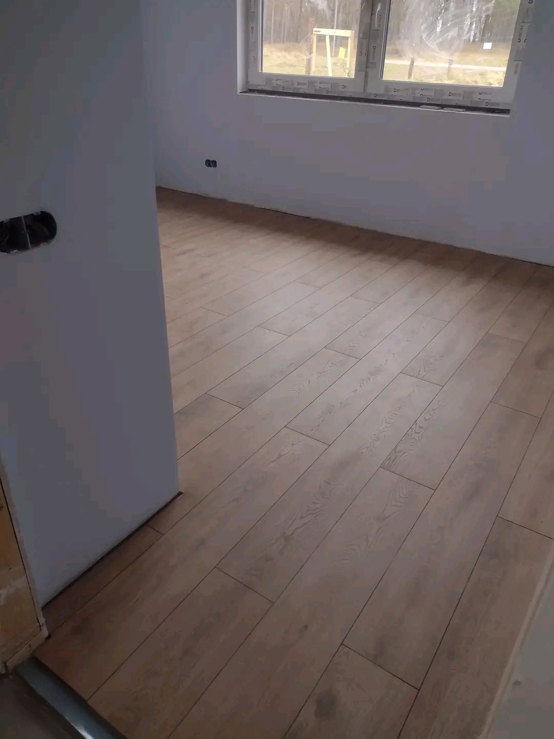 Ułożone panele podłogowe w jasnym odcieniu drewna w nowym pomieszczeniu z oknem, widoczne gniazdka elektryczne i otwór na instalację elektryczną.