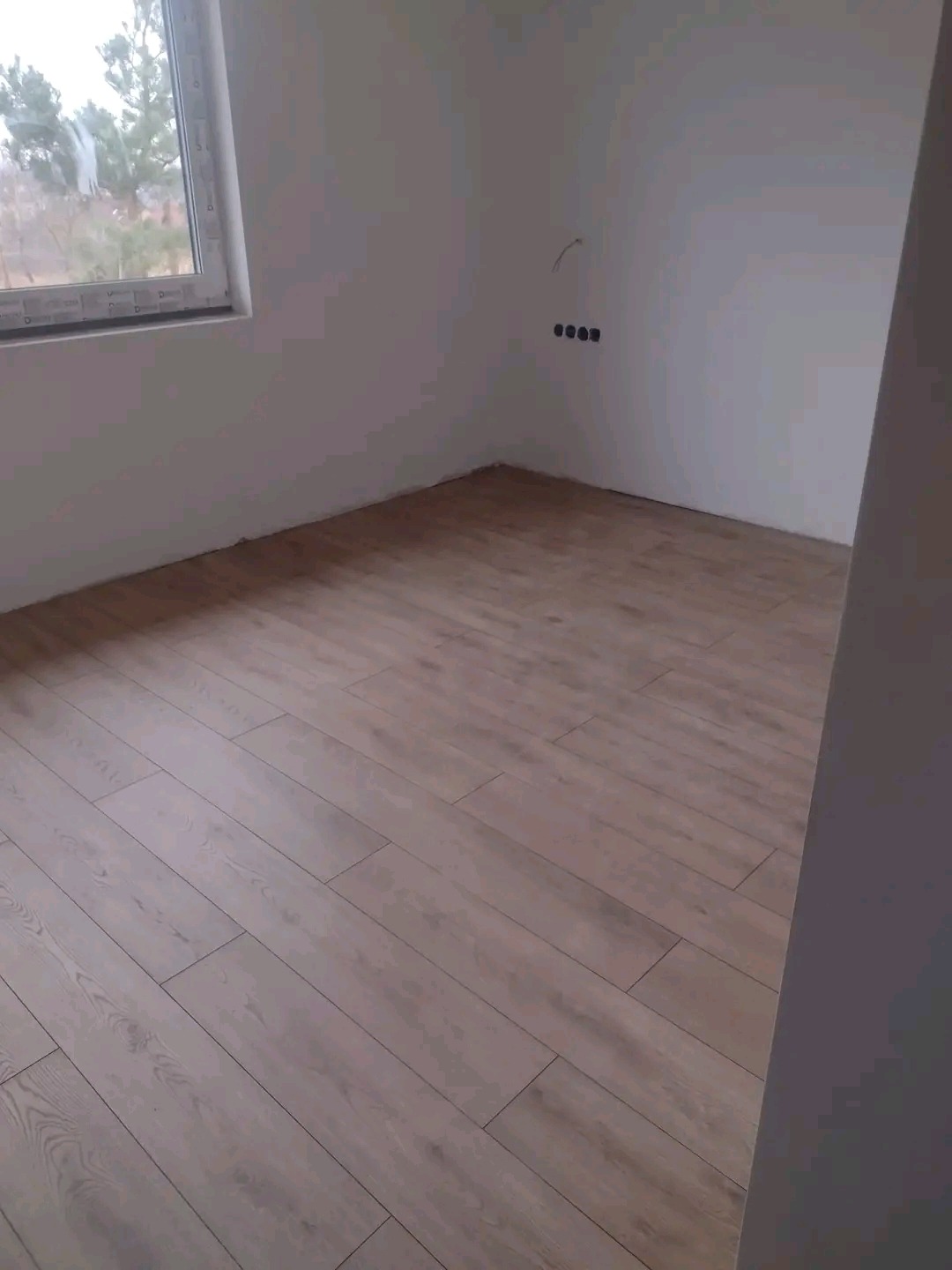 Jasne panele podłogowe ułożone w nowym pokoju z gładkimi, białymi ścianami i oknem z widokiem na drzewa.