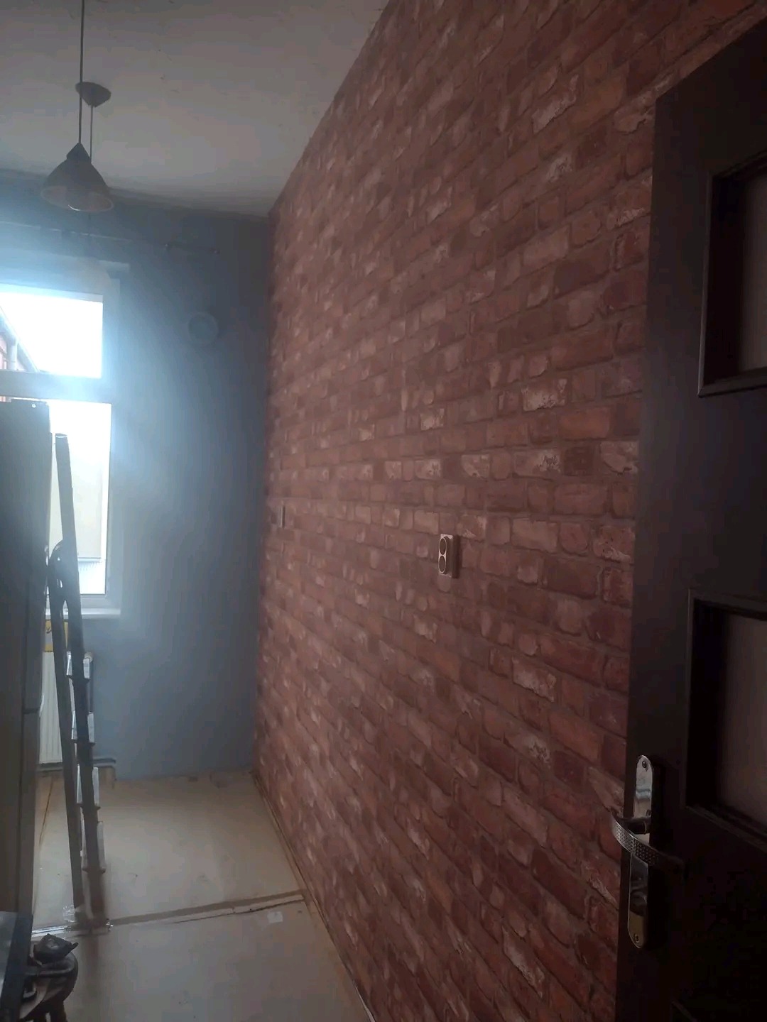 Ściana wykończona imitacją czerwonej cegły, z widocznym gniazdkiem elektrycznym i fragmentem ciemnych drzwi, w tle ściana pomalowana na szaro i drabina oparta o ścianę przy oknie.