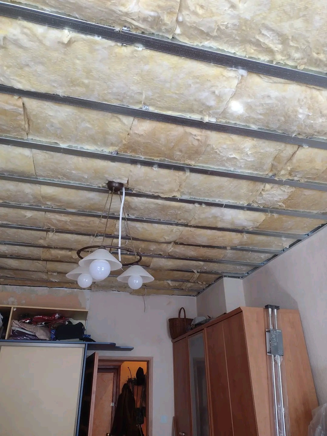 Montaż izolacji z wełny mineralnej na suficie podwieszanym z metalową konstrukcją, widoczna lampa wisząca i fragment umeblowania pokoju.