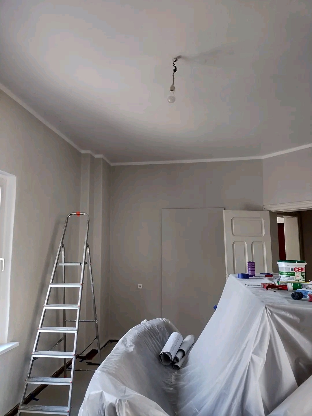 Pokój w trakcie malowania, zabezpieczone meble folią, drabina oparta o ścianę, widoczne wałki malarskie i puszka farby CEKOL.
