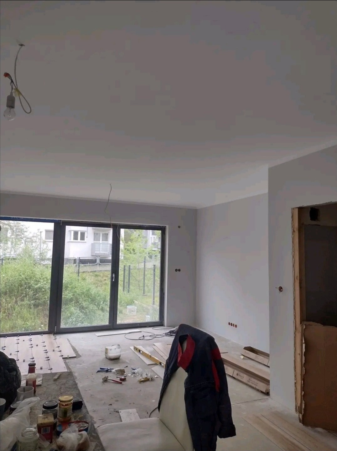 Wnętrze w trakcie prac wykończeniowych: świeżo pomalowane ściany, przygotowane panele podłogowe, narzędzia i odzież robocza na krześle, widok na zieleń za oknem.