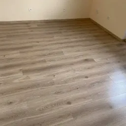 Jasne panele podłogowe imitujące drewno, ułożone w pustym pokoju z kremowymi ścianami i listwami przypodłogowymi w kolorze paneli, widoczne gniazdka elektryczne na ścianie.