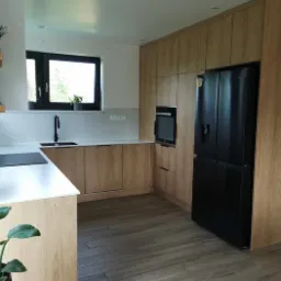 Jasna kuchnia z drewnianymi frontami, czarną lodówką i armaturą, oraz podłogą imitującą drewno, z rośliną doniczkową w lewym dolnym rogu kadru.