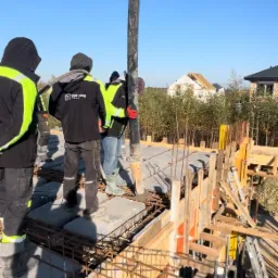 Wylewanie betonu na strop domu murowanego, widoczne zbrojenia i szalunki, w tle dom w stanie surowym otwartym.