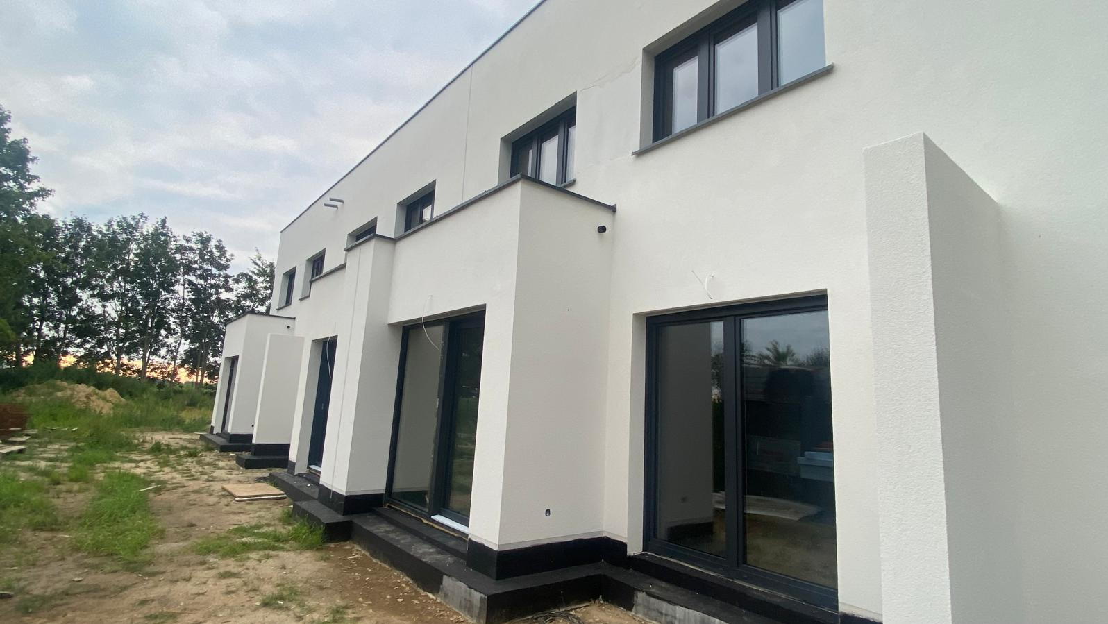 Szeregowy budynek mieszkalny w stanie surowym zamkniętym, z widocznymi czarnymi ramami okiennymi i drzwiowymi oraz fragmentem niezagospodarowanego terenu.