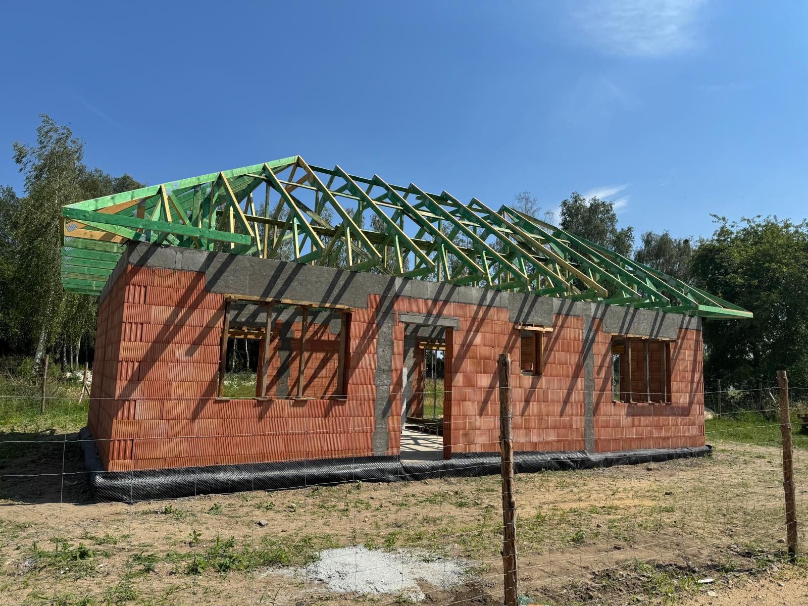 Surowy stan domu z czerwonej cegły z widocznym szkieletem dachu z impregnowanego drewna, otoczony siatką ogrodzeniową i zielenią.