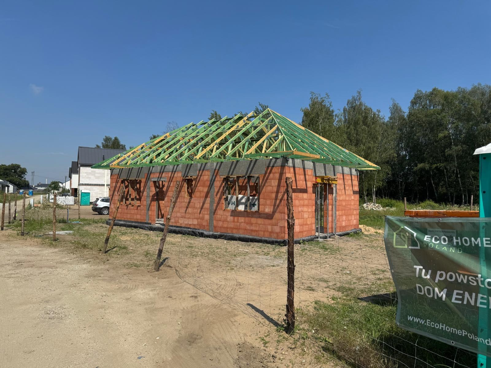 Surowy stan domu z czerwonej cegły z widoczną konstrukcją dachu z zielonych belek, otoczony tymczasowym ogrodzeniem i reklamą firmy budowlanej.