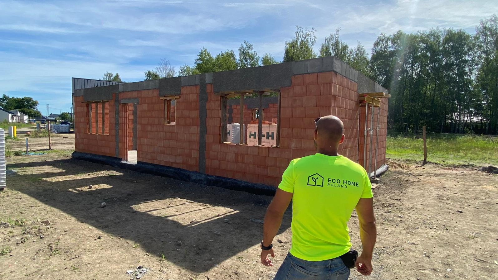 Wznoszenie ścian parterowego domu z czerwonej cegły, widok zza pleców pracownika w neonowej koszulce Eco Home Poland, w tle drzewa i błękitne niebo.