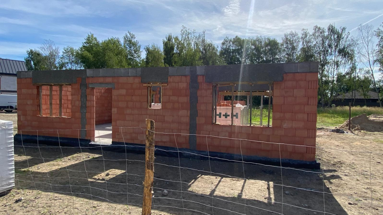 Surowy stan domu murowanego z czerwonej cegły, widoczne otwory okienne i drzwiowe, świeżo wylane nadproża, w tle drzewa i niebo.