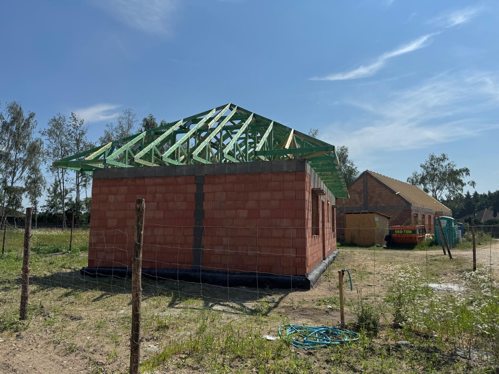 Wznoszenie ścian i konstrukcji dachu domu z czerwonej cegły, widoczne materiały budowlane i teren budowy.