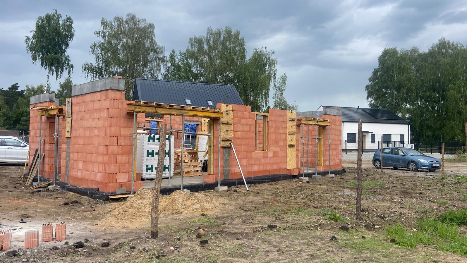 Wznoszenie ścian parterowego domu z czerwonej cegły, widoczne drewniane szalunki okienne i drzwiowe, palety z materiałem budowlanym, w tle dom jednorodzinny i drzewa.