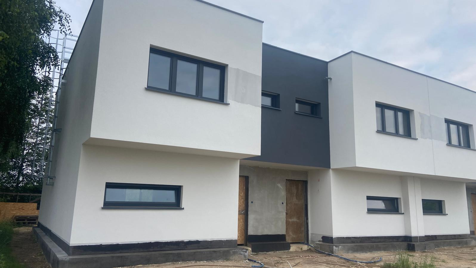 Nowoczesne bliźniaki w trakcie wykończenia elewacji, z widocznymi surowymi drzwiami wejściowymi i rusztowaniem po lewej stronie.