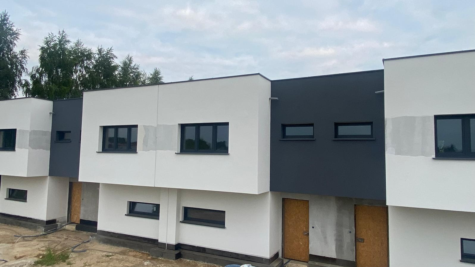 Nowoczesne domy szeregowe w trakcie wykończenia elewacji, z widocznymi śladami po szpachlowaniu i zamontowanymi oknami w kolorze antracytowym, z tymczasowymi drzwiami wejściowymi.