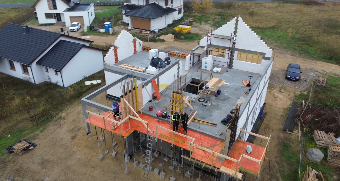 Wysoka perspektywa na plac budowy z dwoma budynkami w stanie surowym otwartym, widoczni pracownicy w odblaskowych kamizelkach, szalunki i podpory, w tle zabudowa jednorodzinna i jesienne drzewa.