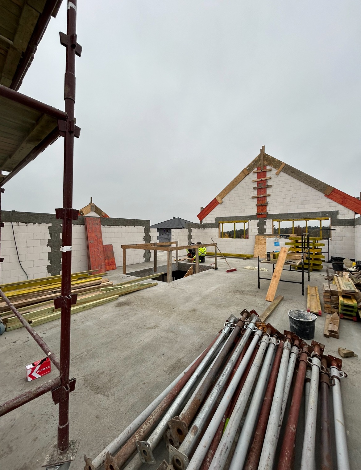 Budowa domu z betonu komórkowego w trakcie realizacji, widoczne rusztowania, podpory i materiały budowlane, pracownik w odblaskowej kamizelce przy otworze w stropie.