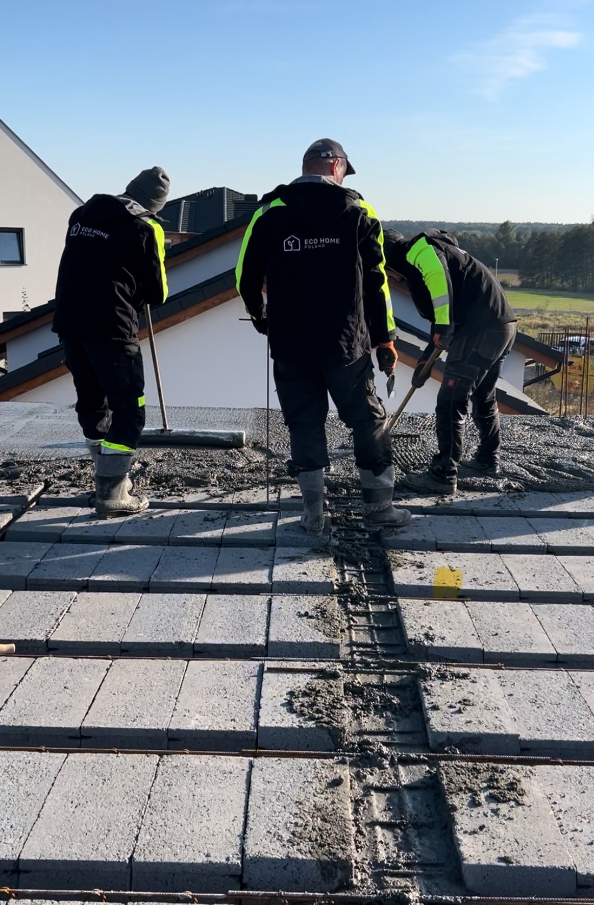 Trzech pracowników w odzieży roboczej z logo firmy 'Eco Home Poland' układa szare bloczki na dachu domu, widoczna siatka zbrojeniowa i zaprawa, w tle krajobraz.