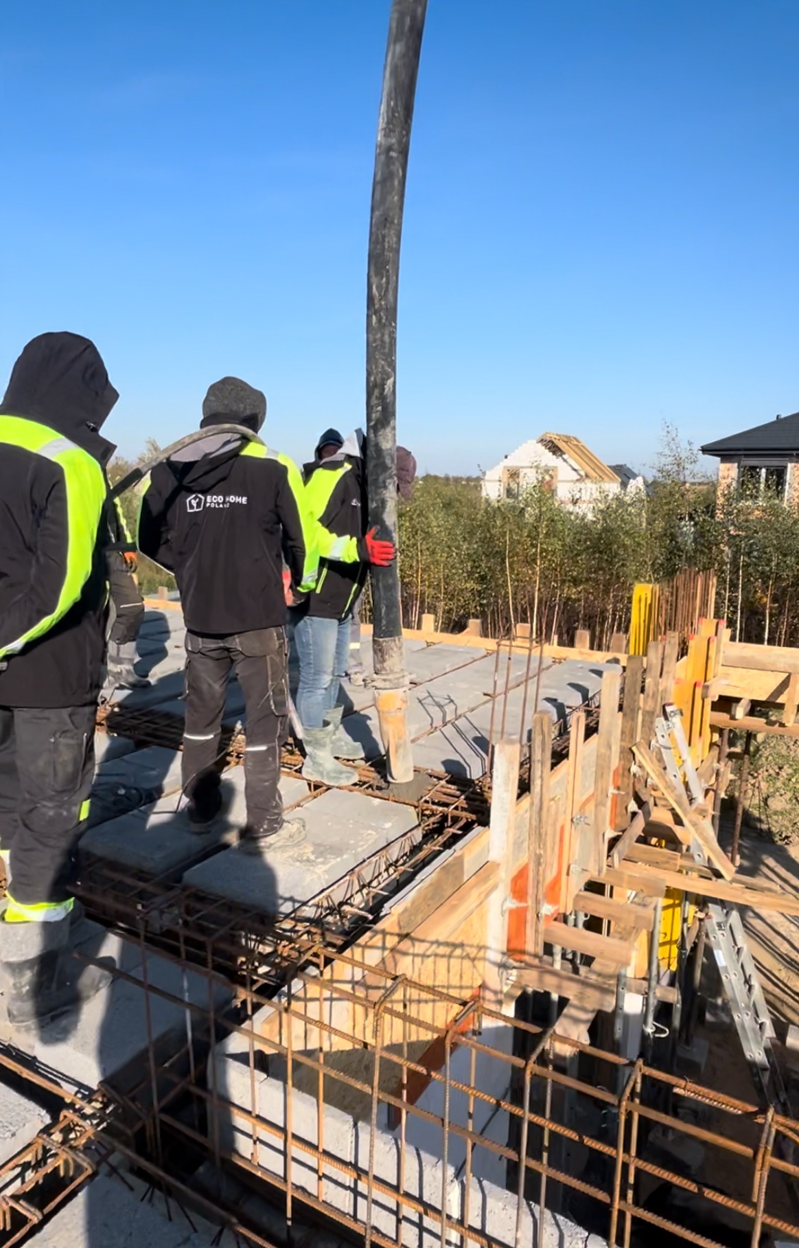 Wylewanie betonu na strop domu murowanego, widoczne zbrojenia i szalunki, w tle dom w stanie surowym otwartym.