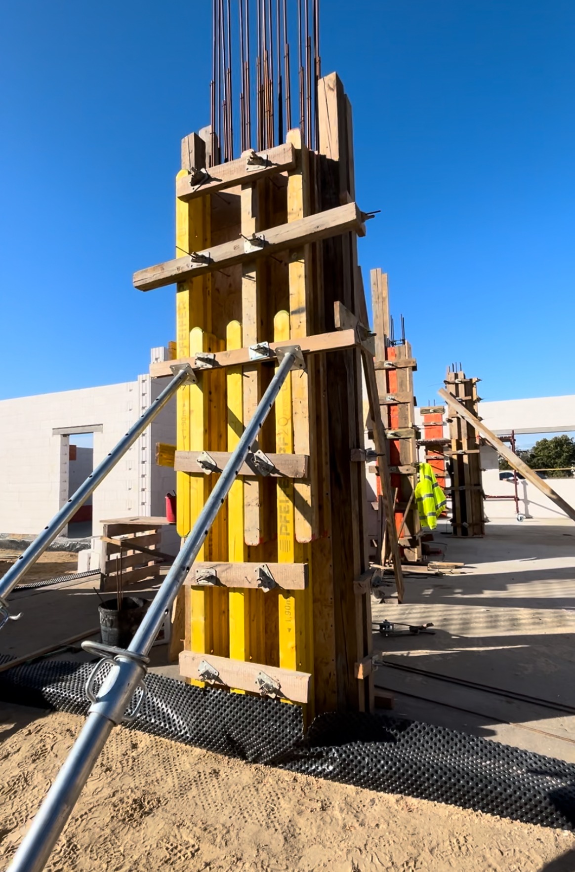 Szare metalowe podpory stabilizują szalunek kolumny z żółtymi panelami i drewnianymi belkami na placu budowy, widoczny pracownik w odblaskowej kamizelce oraz fragment budynku z pustaków.