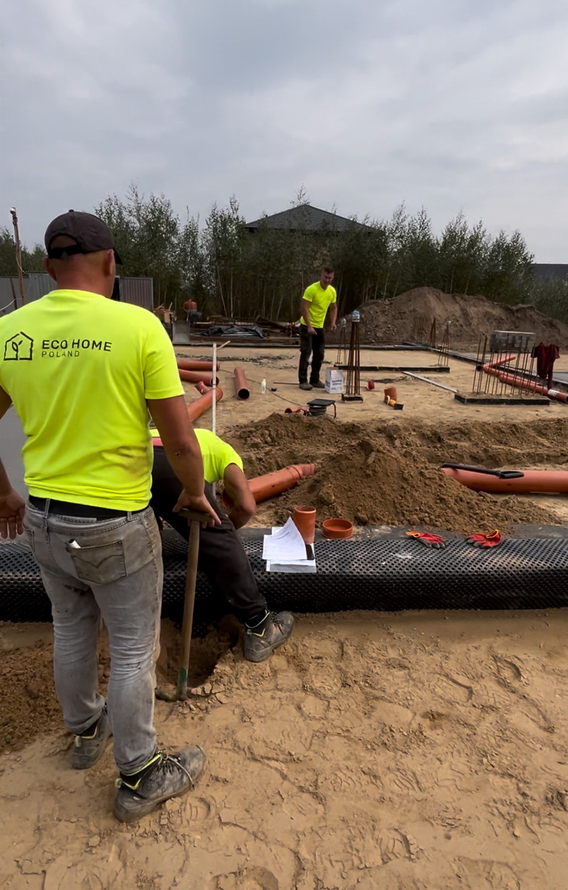 Prace ziemne przy budowie domu: robotnicy w jaskrawych koszulkach z logo firmy układają rury kanalizacyjne na czarnej folii izolacyjnej, widoczne zbrojenia fundamentów i kopiec ziemi w tle.