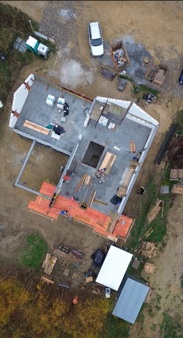 Widok z góry na plac budowy domu jednorodzinnego w trakcie wznoszenia ścian, widoczne fundamenty, częściowo ułożone ściany parteru, rusztowania, materiały budowlane i samochód na działce.