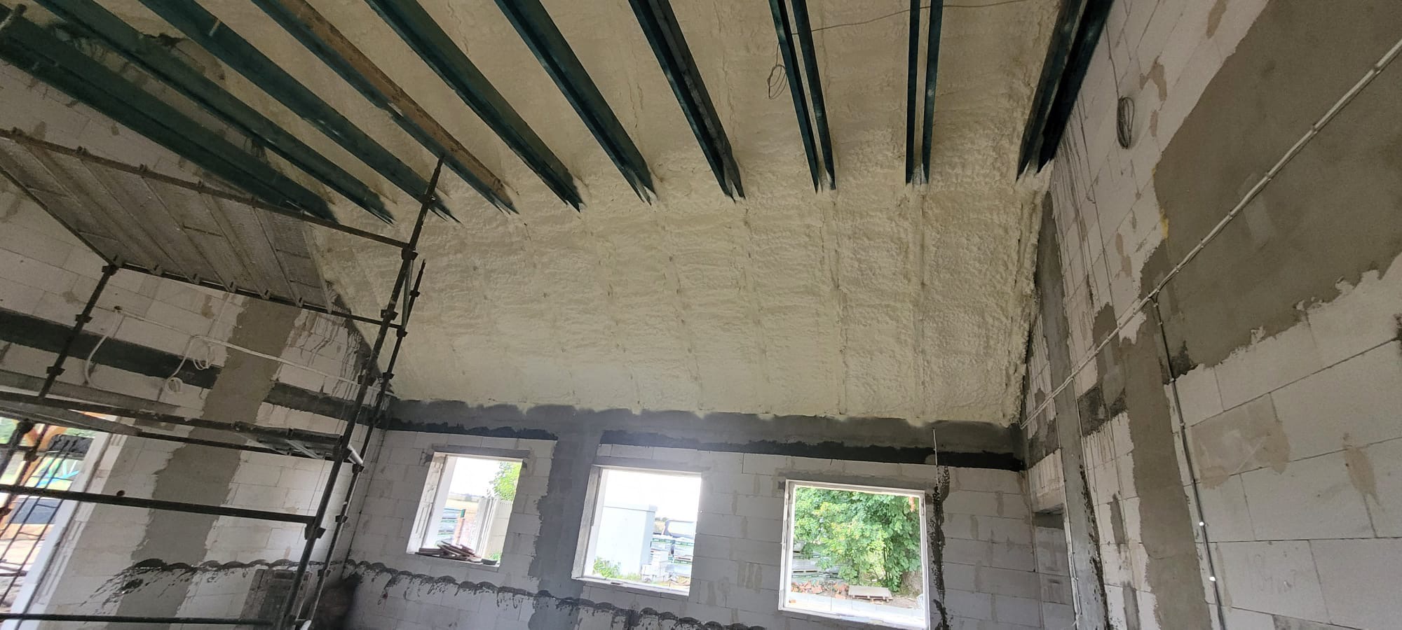 Wnętrze w trakcie ocieplania pianką na poddaszu, widoczne metalowe belki stropowe i ściany z bloczków betonowych, częściowo otynkowane, z trzema oknami z widokiem na zieleń, oraz rusztowanie...