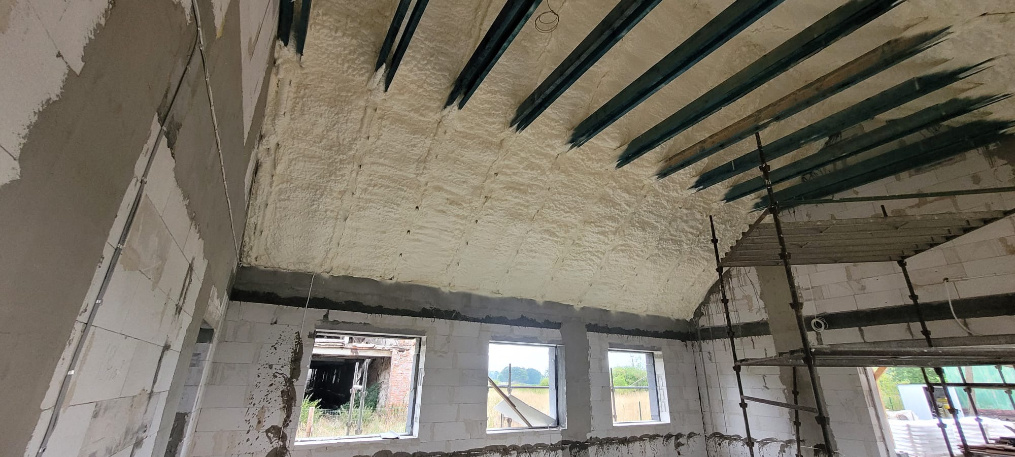 Poddasze domu w trakcie ocieplania pianką PUR, widoczne metalowe belki stropowe i rusztowanie, surowe ściany z betonu komórkowego i okna wychodzące na zewnątrz.