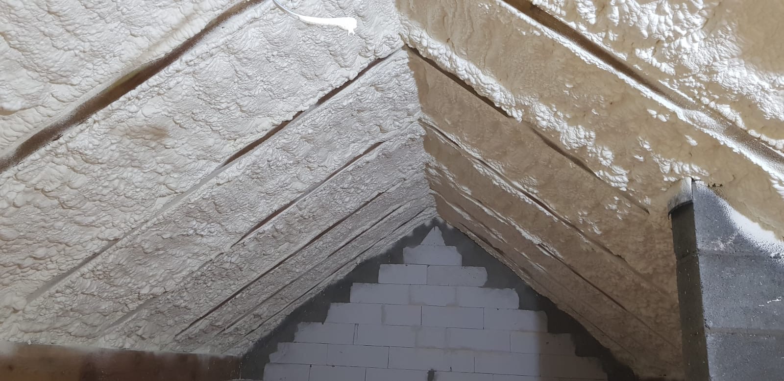 Poddasze z widoczną konstrukcją drewnianą i warstwą izolacyjną z pianki, z fragmentem ściany z białych bloczków betonowych u dołu, w tle komin