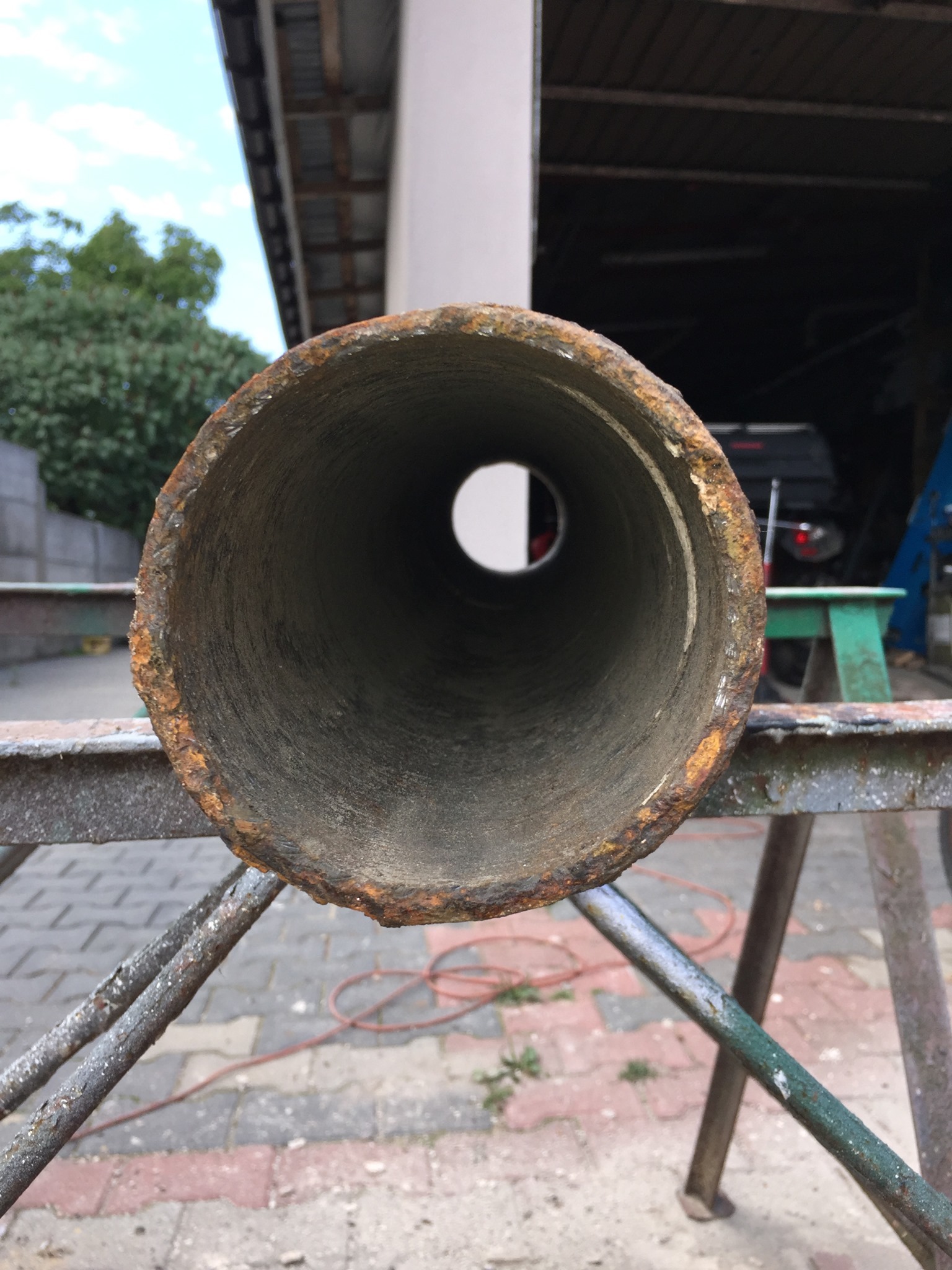 Wnętrze skorodowanej rury kanalizacyjnej, widok z perspektywy wewnątrz, na tle warsztatu i zieleni.