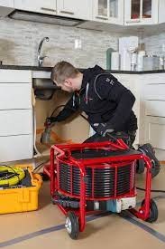 Hydraulik w trakcie pracy pod zlewem, używa specjalistycznej maszyny z czerwonym stelażem i czarnym wężem do czyszczenia rur. W tle widoczne białe szafki kuchenne i bateria.