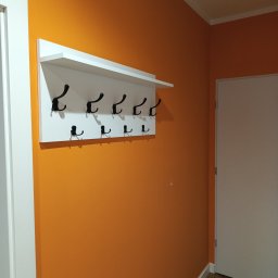 Michał  Wojtas - Pomarańczowa ściana z białym wieszakiem na ubrania z czarnymi haczykami. Widoczny fragment białych drzwi i podłogi.