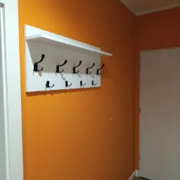 Pomarańczowa ściana z białym wieszakiem na ubrania z czarnymi haczykami. Widoczny fragment białych drzwi i podłogi.