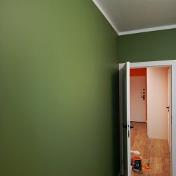 Michał  Wojtas - Ściana pomalowana na oliwkowy kolor z otwartymi drzwiami, przez które widać podłogę i akcesoria malarskie. Widoczny fragment sufitu i listwy przypodłogowe.