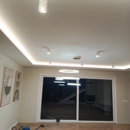 Michał  Wojtas - Jasne wnętrze po remoncie z parkietem w jodełkę. Widoczny sufit z oświetleniem LED, nowoczesne lampy wiszące i odkurzacz. Minimalistyczny design.