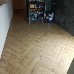 Michał  Wojtas - Ułożona podłoga z paneli w jodełkę, widoczny wózek z narzędziami i fragment ściany z czarnymi płytkami. W tle widoczne kolejne pomieszczenie.