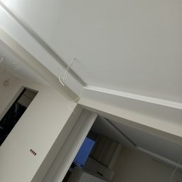 Michał  Wojtas - Biały sufit po malowaniu z listwami dekoracyjnymi, widoczne wystające kable i fragment ściany z gniazdkami. Ujęcie z dołu, perspektywa wnętrza w trakcie wykończenia.