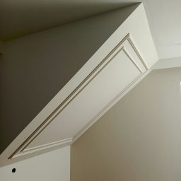 Michał  Wojtas - Wykończone poddasze ze skosami, pomalowane na biało z ozdobnymi listwami. Widoczny fragment drewnianej podłogi i otwór na gniazdko elektryczne.