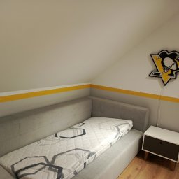 Michał  Wojtas - Pokój na poddaszu z szarą sofą i żółtym pasem na ścianie, nad którym wisi lampka z logiem drużyny hokejowej Pittsburgh Penguins. Minimalistyczny styl.