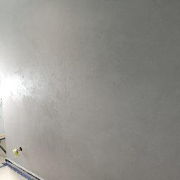 Michał  Wojtas - Ściana po gładzi w odcieniach szarości, zabezpieczona podłoga folią malarską. Widoczne delikatne refleksy światła na powierzchni ściany. Gniazdko elektryczne zabezpieczone taśmą.